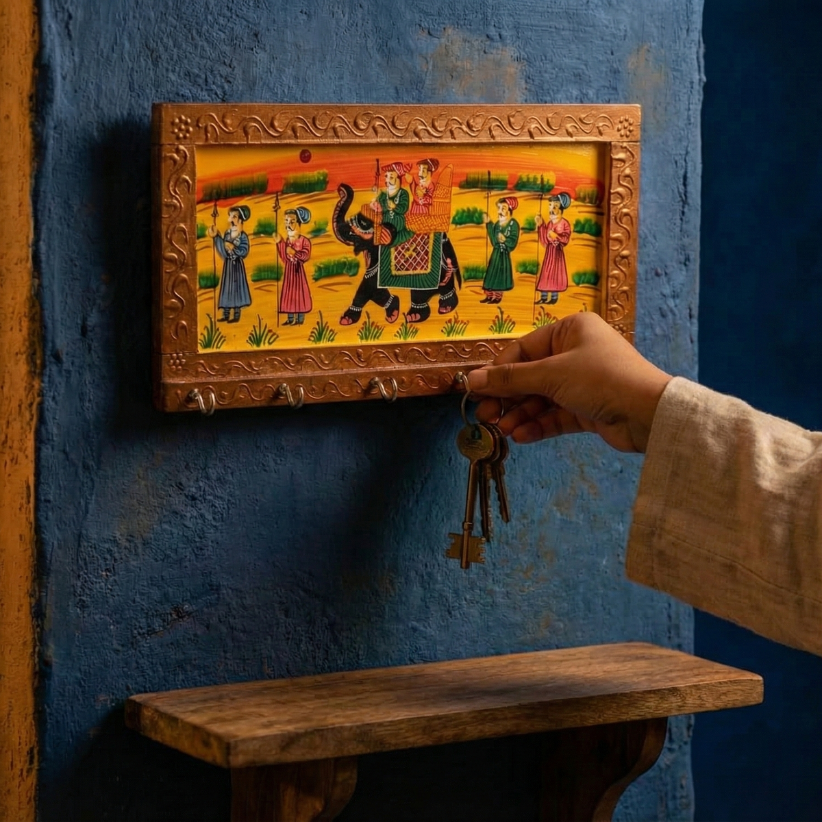 A hand hangs keys from a handcrafted golden brown wooden key holder for the wall featuring traditional Rajasthani elephant painting, hanging in an entryway surrounded by green plants.