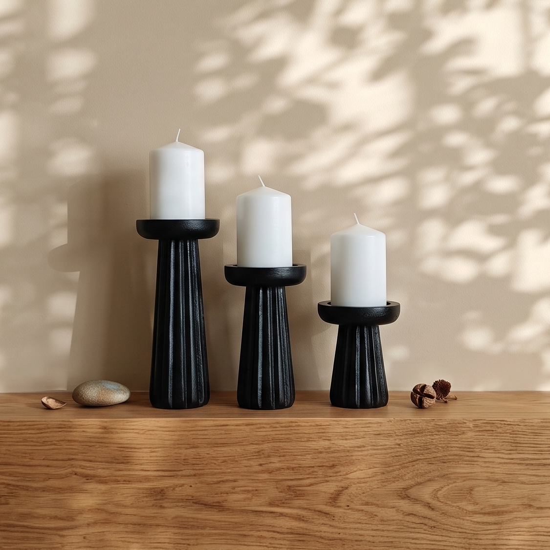 Three black fluted pillar candle holders with white candles styled on a thick wooden fireplace mantel, surrounded by dappled sunlight and leaf shadows on a neutral beige wall.