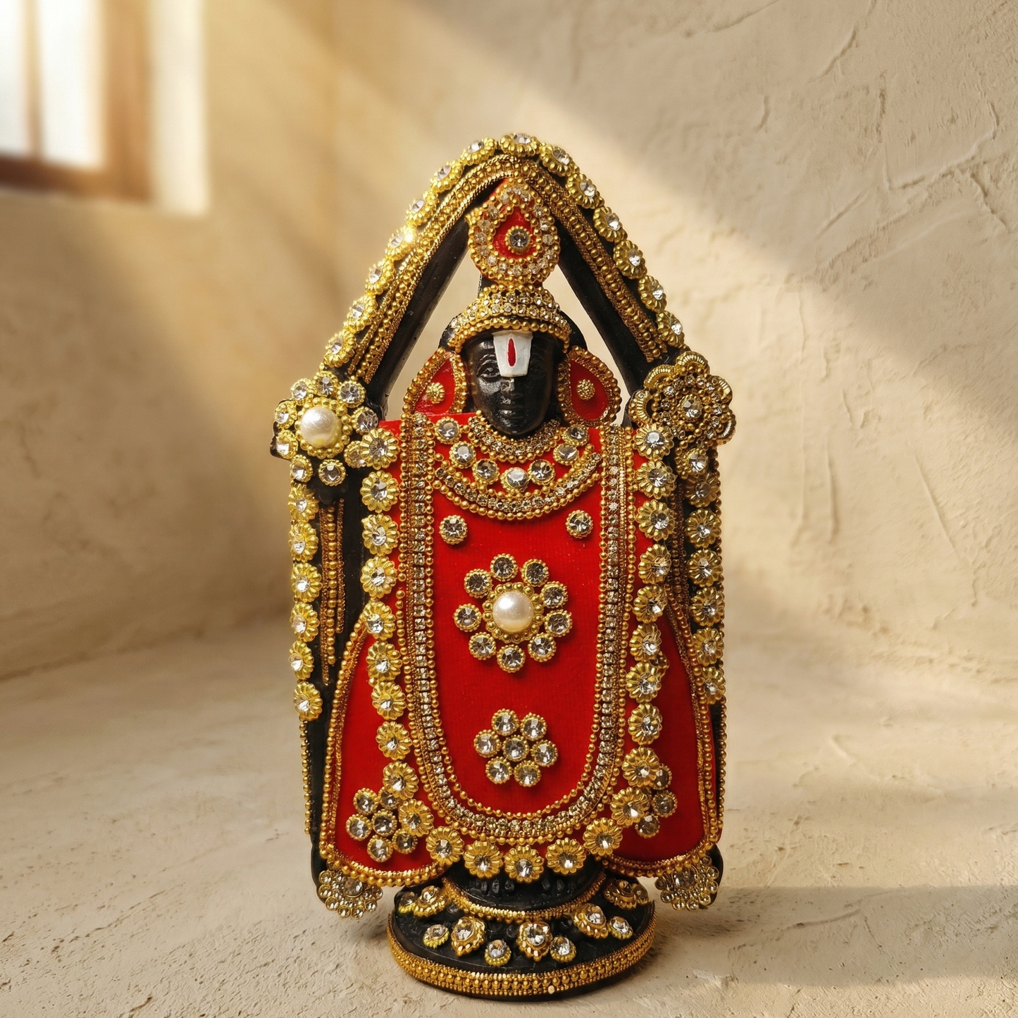 Handcrafted black Lord Venkateswara (Tirupati Balaji) murti richly adorned with red fabric, gold-tone detailing, and sparkling stones with a beam of sunlight on the lord's face