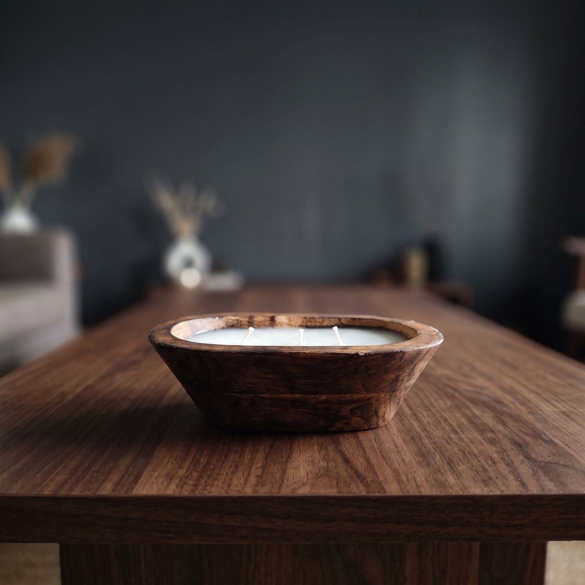 A solid natural wood oval dough bowl candle with three unlit wicks, sitting as a natural wood centerpiece on a dark walnut coffee table in a softly blurred, modern moody living room.