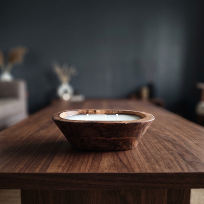 A solid natural wood oval dough bowl candle with three unlit wicks, sitting as a natural wood centerpiece on a dark walnut coffee table in a softly blurred, modern moody living room.