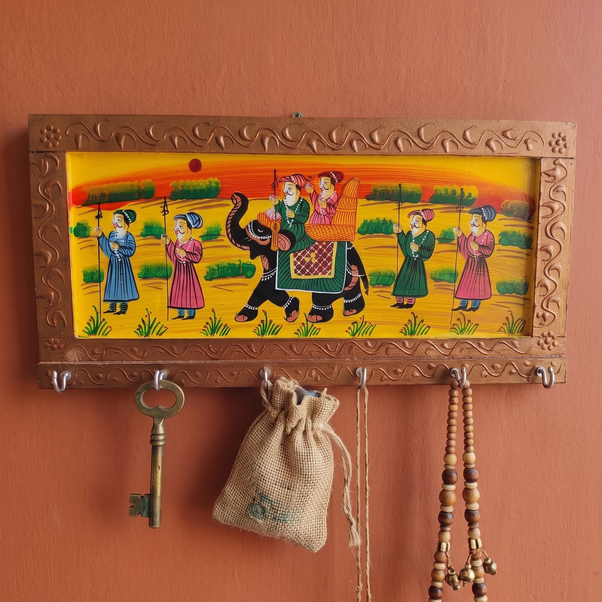 A handcrafted golden brown wooden key holder for the wall featuring traditional Rajasthani elephant painting, hanging in an entryway surrounded by green plants.