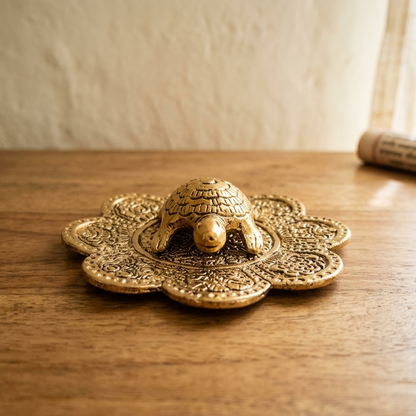 Eye-level view of a highly detailed aluminum tortoise with gold polish on a carved floral stand, resting on a warm wooden surface next to a rolled manuscript in soft golden hour light.