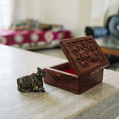A small metal Nandiji figurine with a dark antique finish sitting on a light stone table next to a small, open carved wooden box with a red interior.
