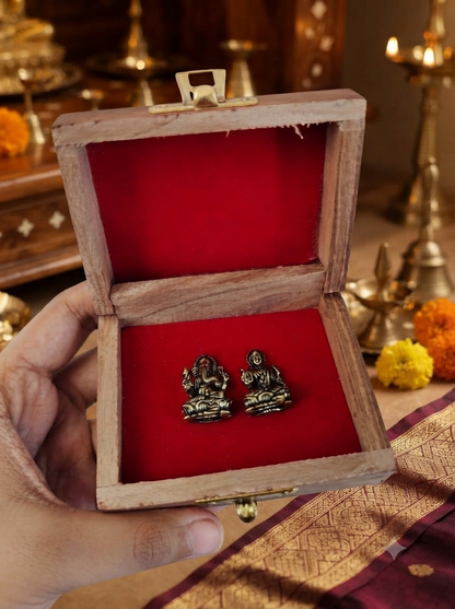 A person holding open a small wooden box lined with bright red velvet, revealing miniature antique finish metal Laxmi and Ganesha idols nestled inside, with traditional oil lamps in the background.