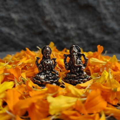 A pair of small, detailed antique finish metal Laxmi and Ganesha idols resting on a bed of vibrant yellow and orange marigold petals against a dark stone background.
