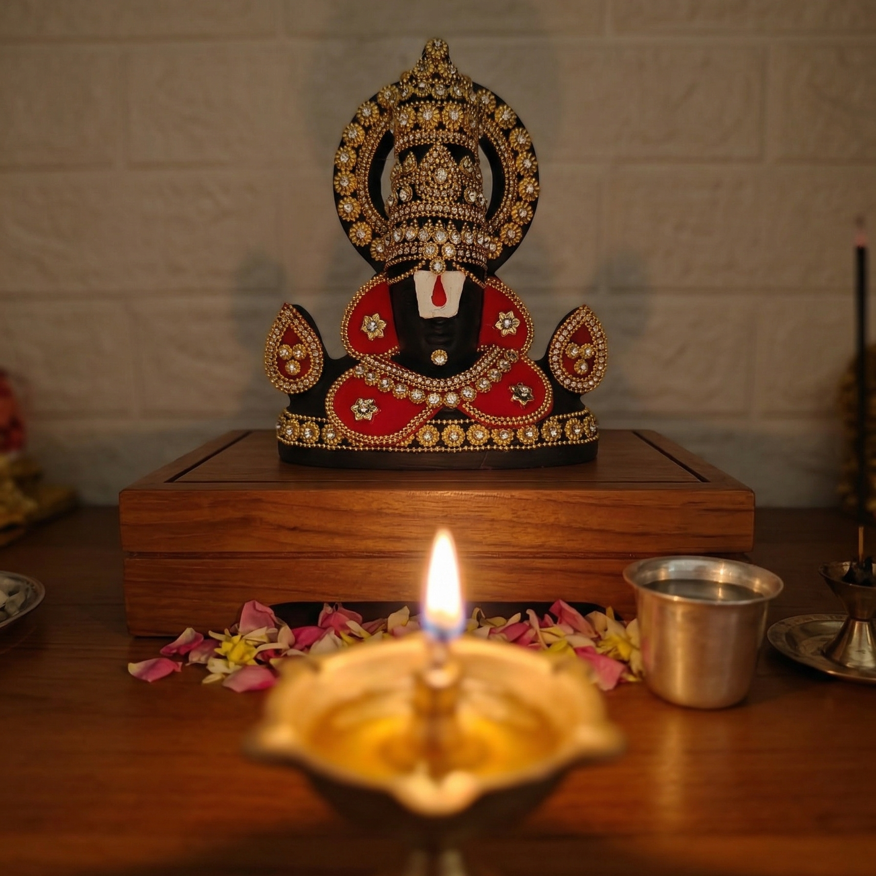 Black Tirupati Balaji divine face adorned with red and gold-tone stone embellishments, surrounded by flower petals and a lit diya.
