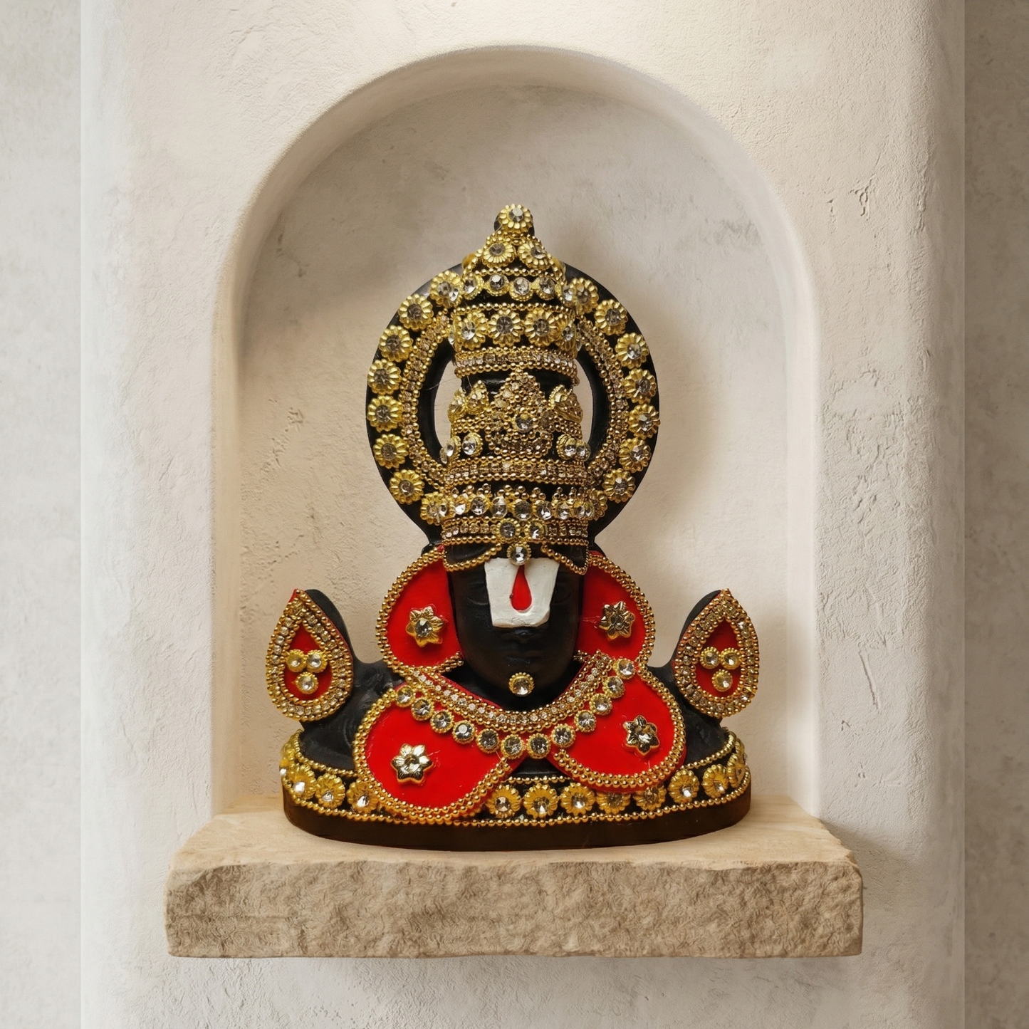 Black Tirupati Balaji divine face adorned with red and gold-tone stone embellishments against a light gray wall