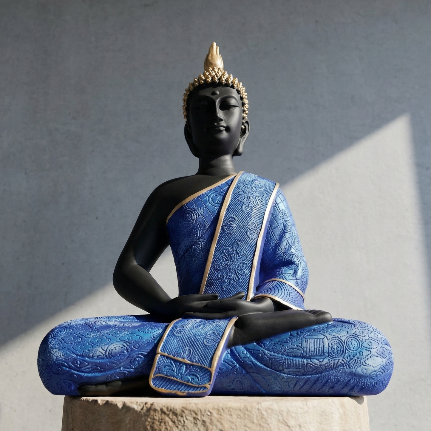 Black meditating Buddha statue in Dhyana mudra wearing intricately textured blue robe with gold borders against a gray background