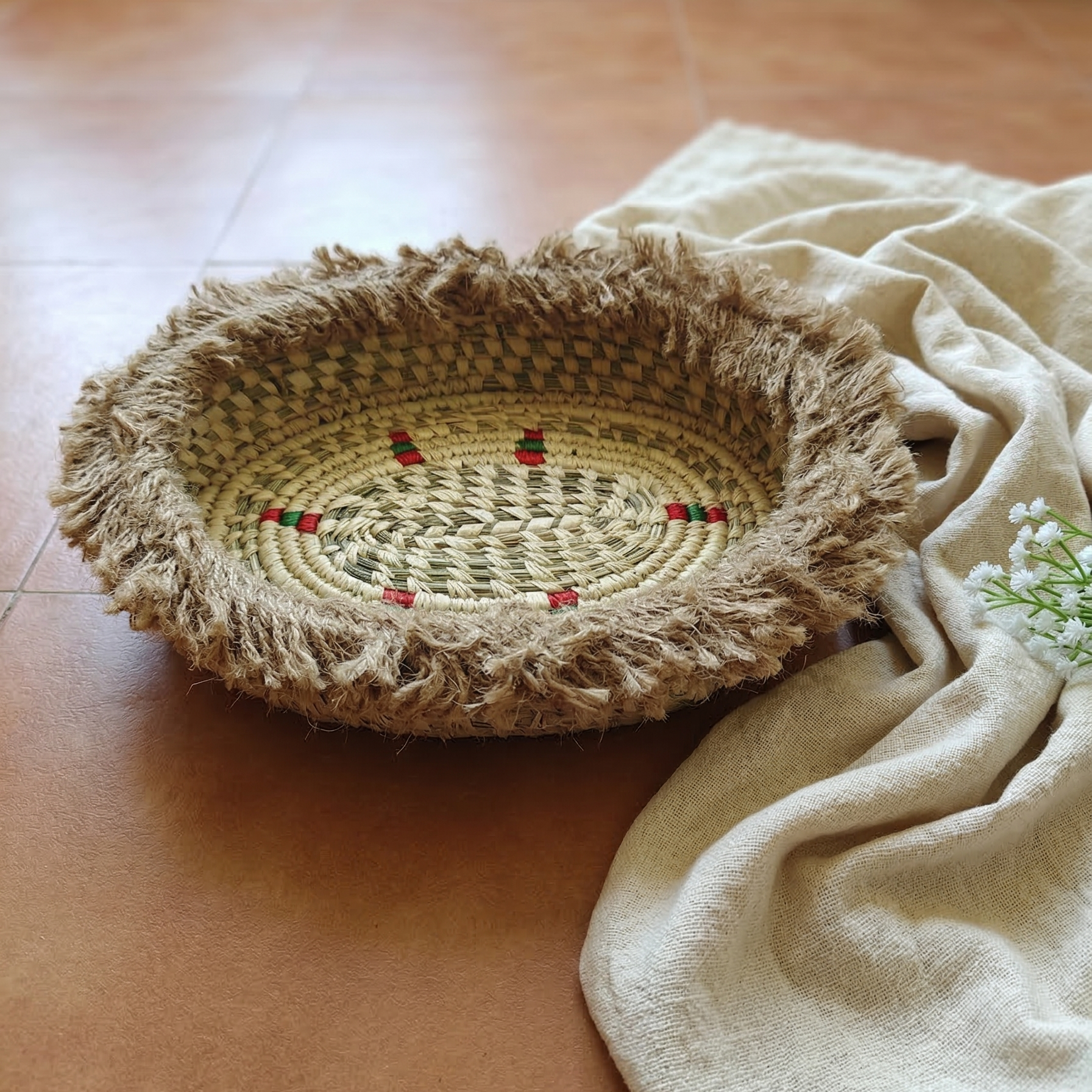 A close-up photograph of an oval, handwoven sabai grass basket featuring a raw, shaggy fringe and subtle red and green woven accents. It is resting on a warm, terracotta tiled floor next to a softly draped cream cloth.