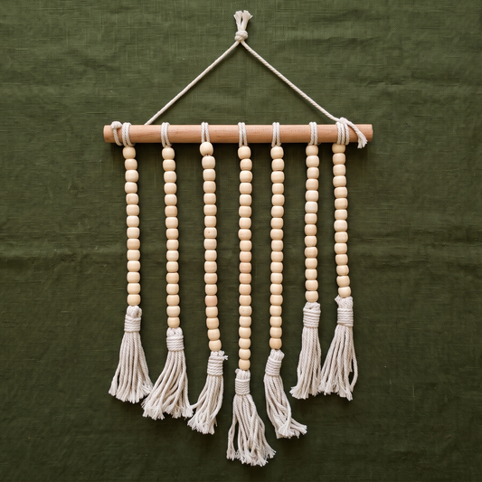 A boho-style wall hanging featuring seven strands of natural wooden beads ending in cotton fringe tassels, suspended from a wooden dowel against a dark olive green fabric background.