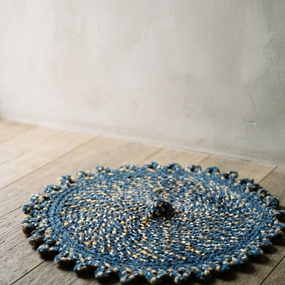 A circular braided rug in shades of coastal blue and beige with a looped decorative border, resting on a wide plank wooden floor beneath a simple black wall hook.