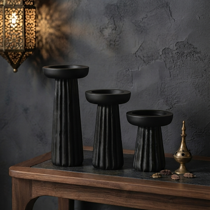 A set of three empty black fluted candle stands resting on a dark slate-top console table, styled in a moody room with a glowing ornate brass lantern hanging in the background.