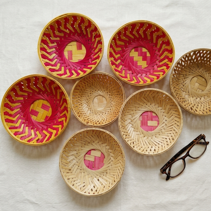 Top-down view of seven woven bamboo bowls in natural and vibrant pink patterns resting on white linen, with tortoiseshell glasses placed nearby for scale.