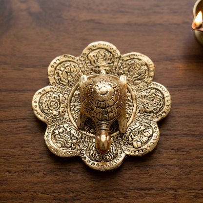 op-down view of a symbolic aluminum turtle with a shiny gold finish resting on an ornate floral metal base, displayed on a rich dark wood table near a glowing oil lamp.