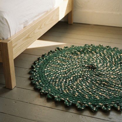 A deep green circular braided rug with a textured scalloped border placed on grey painted floorboards next to a light oak wooden bed frame with crisp white sheets.