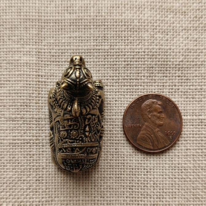 A highly detailed, miniature metal Nandiji figurine with a darkened antique finish placed directly next to a US penny for scale on a textured beige fabric.