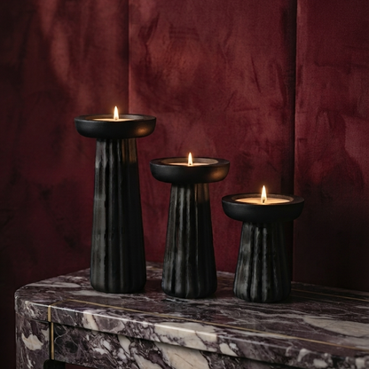 Three lit tealight candles placed in black carved wooden holders of descending heights, sitting on a luxurious purple marble console table against a deep burgundy velvet wall.