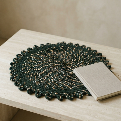 A round, woven emerald green mat with a scalloped edge and subtle gold flecks, styled on a smooth beige stone table with a simple linen-bound notebook resting on top.