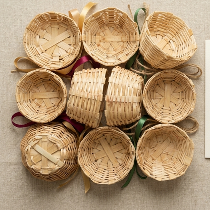 Ten small round woven bamboo baskets are meticulously arranged into a square pattern on a textured neutral linen surface, interwoven with colorful gold, burgundy, and green ribbons, and a matching jute twine bow, beside a cream-colored product booklet featuring gold text.
