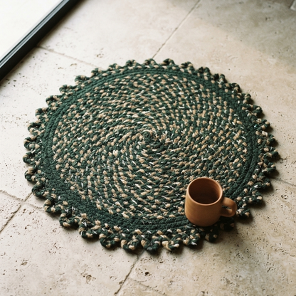 A round, forest green braided rug with natural fiber flecks and a scalloped edge, lying on light travertine stone tiles beside an empty terracotta mug near a sliding glass door.