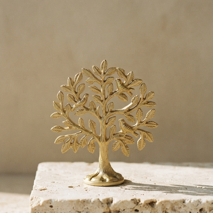 A detailed gold-polished aluminum tree of life resting on a block of porous travertine stone against a smooth, neutral beige background in bright sunlight.