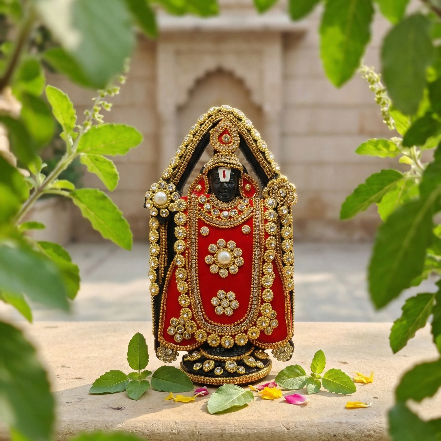 Handcrafted black Lord Venkateswara (Tirupati Balaji) murti richly adorned with red fabric, gold-tone detailing, and sparkling stones with Tulsi Plant in the Background