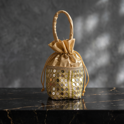 Single golden fabric drawstring potli bag adorned with traditional circular mirror-work, displayed on a marble with a grey background