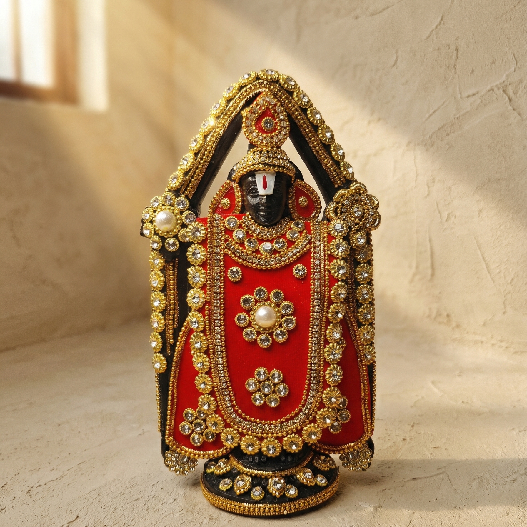 Handcrafted black Lord Venkateswara (Tirupati Balaji) murti richly adorned with red fabric, gold-tone detailing, and sparkling stones with a beam of sunlight on the lord's face