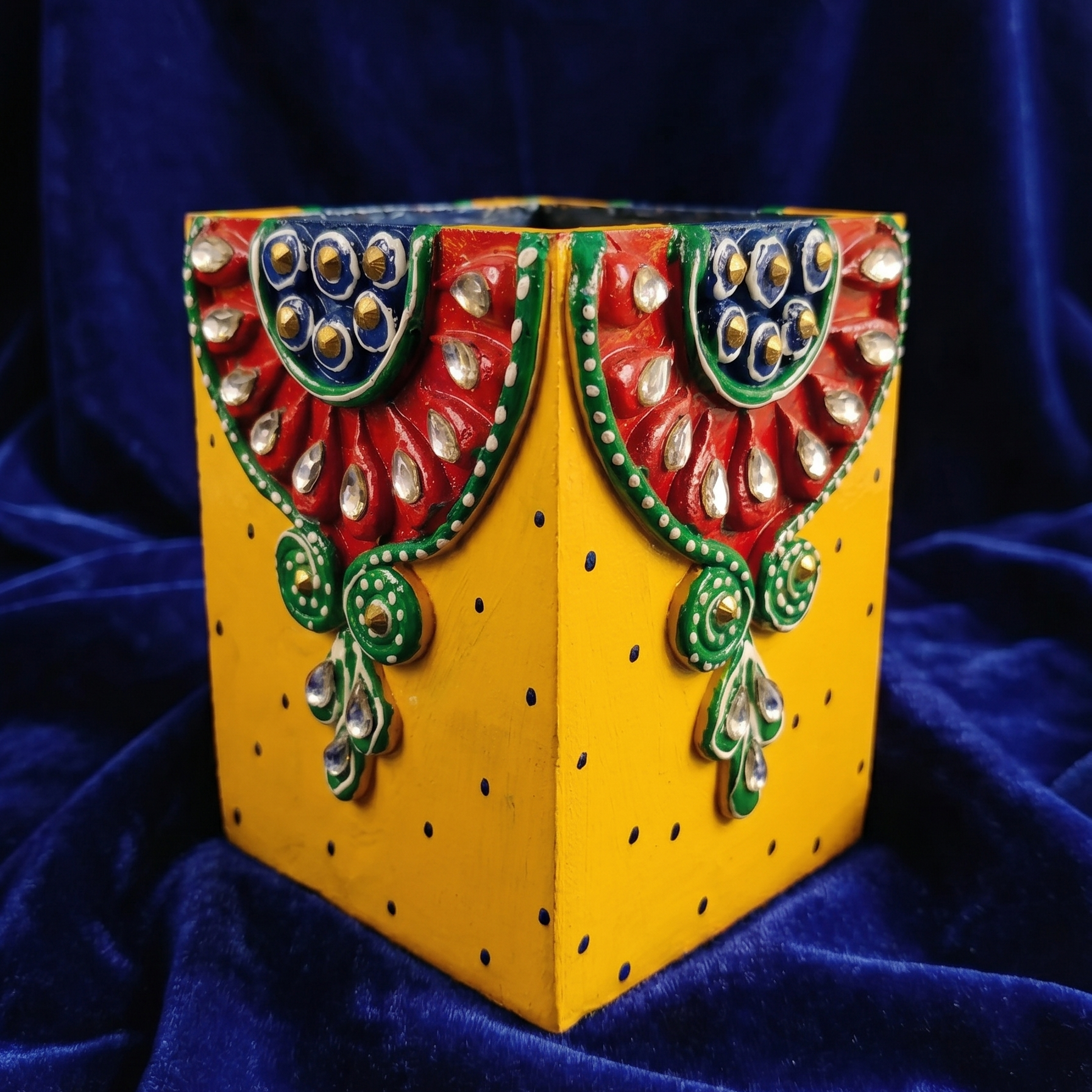 Handcrafted wooden pen holder in bright yellow, decorated with traditional red, green, and blue Kundan mirror work patterns and small polka dots, on a blue background.