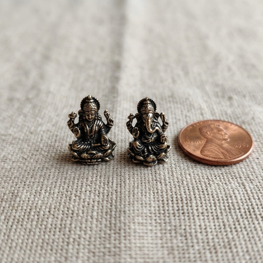 A pair of small dark metallic Ganesha stud earrings shown next to a penny on a textured beige fabric background.