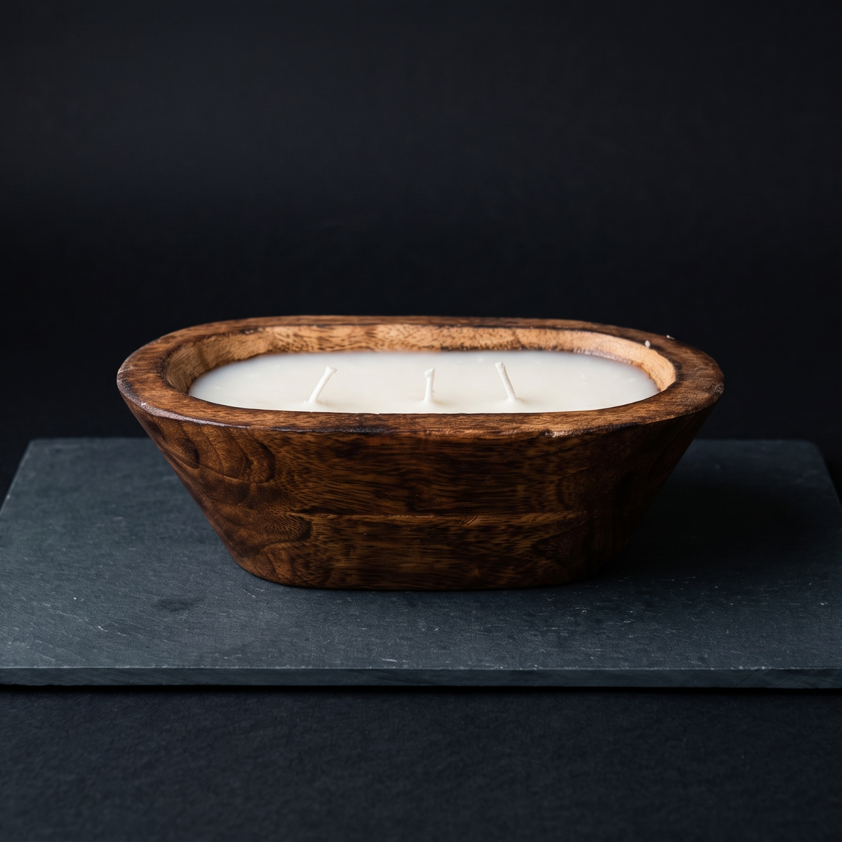 A hand-carved natural wood dough bowl candle featuring three unlit cotton wicks and premium white wax, resting on a dark slate surface against a black background.