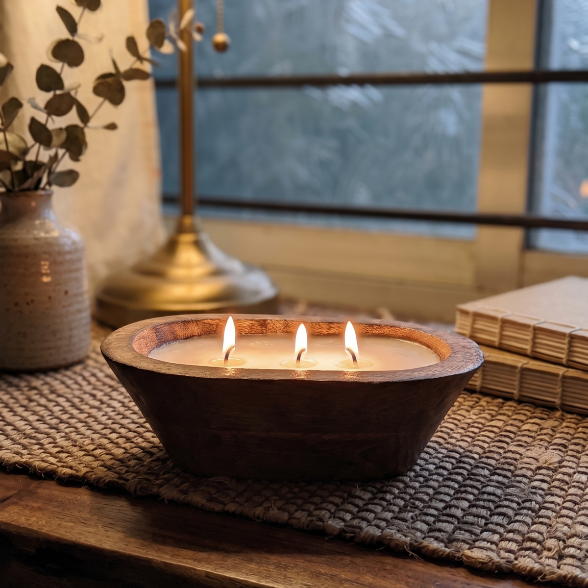 A lit three-wick dough bowl candle with a beautiful pool of melted wax and firelight, styled on a woven mat next to a brass lamp, small vase, and stacked journals in a cozy room.