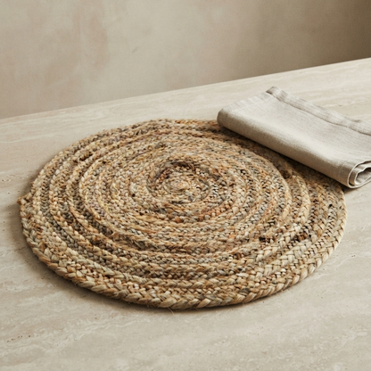 Close-up of a thick, reversible round braided jute placemat resting on a light beige stone surface, paired with a folded natural linen napkin for an earthy table setting.