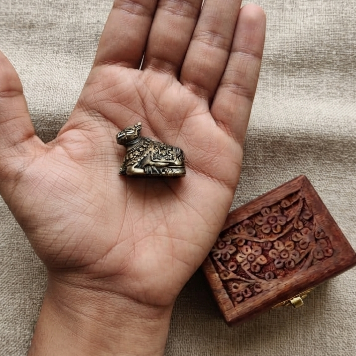 Top-down view of a tiny antique finish metal Nandi resting in the palm of a hand to show scale, placed next to a small carved wooden box on a linen cloth.