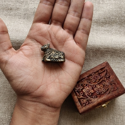 Top-down view of a tiny antique finish metal Nandi resting in the palm of a hand to show scale, placed next to a small carved wooden box on a linen cloth.