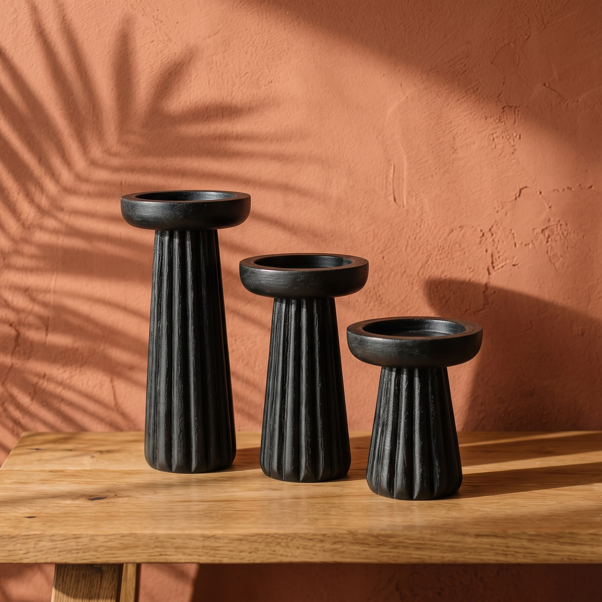 Three empty black ribbed candle pedestals of different heights placed on a natural oak wood table, illuminated by warm sunlight casting palm shadows on a terracotta wall.