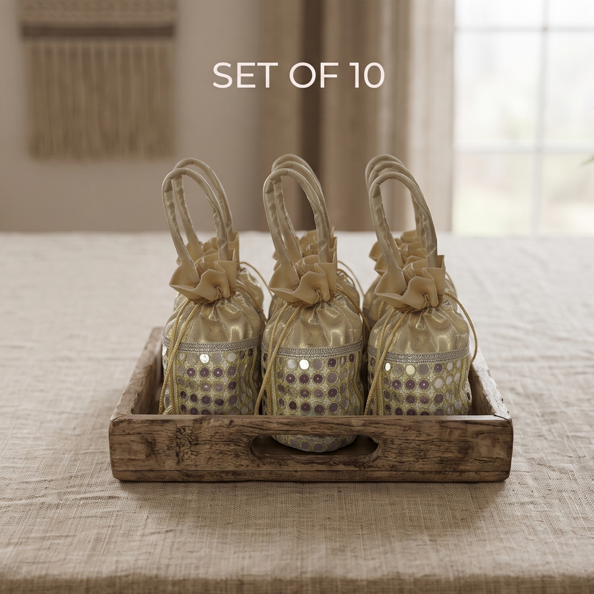 A square wooden tray holding ten golden drawstring pouches with hand-stitched mirror-work, set up as premium party favors on a textured linen tablecloth.