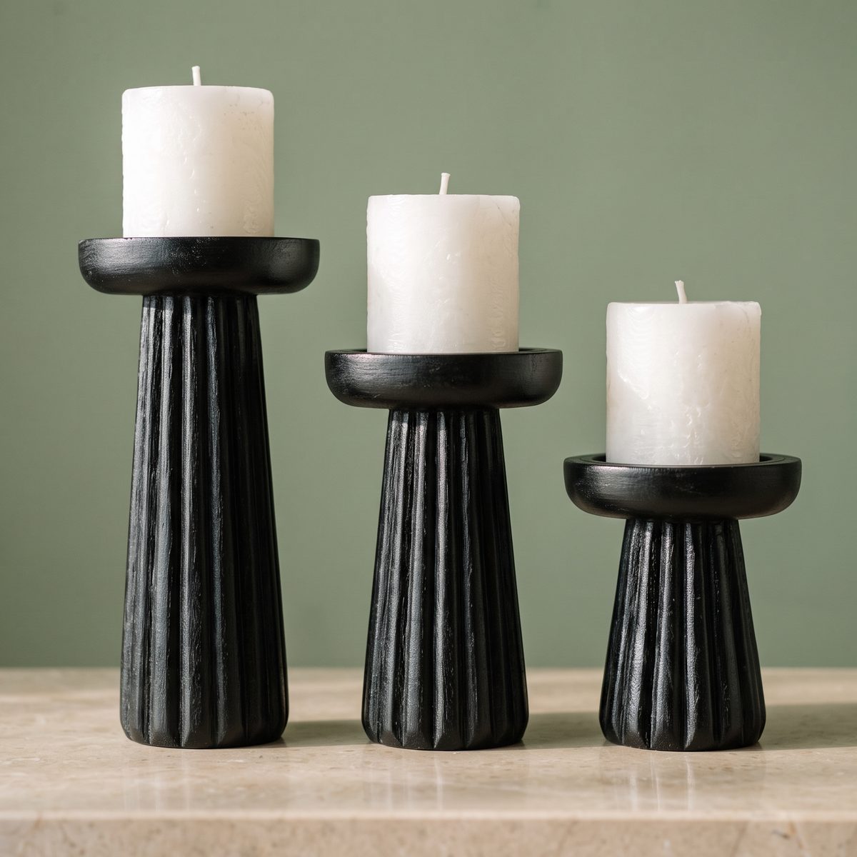 A trio of matte black fluted candle holders carrying chunky white pillar candles, set on a beige stone surface in front of a soft sage green wall.