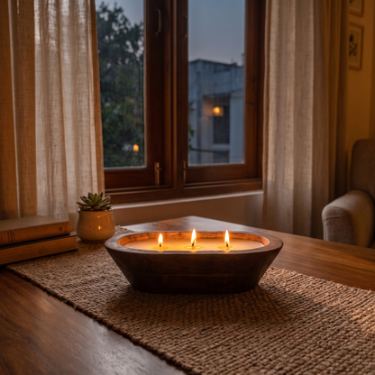 A lit three-wick wooden dough bowl candle casting a warm room-filling glow as a dining table centerpiece, sitting on a woven runner surrounded by books and a succulent near a window at dusk.