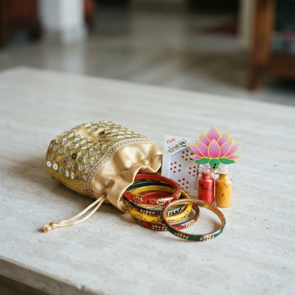A golden mirror-work potli bag gently spilling out vibrant lac bangles, alongside a bindi card and two tiny glass vials of haldi and kumkum topped with a paper lotus motif on a light stone table.