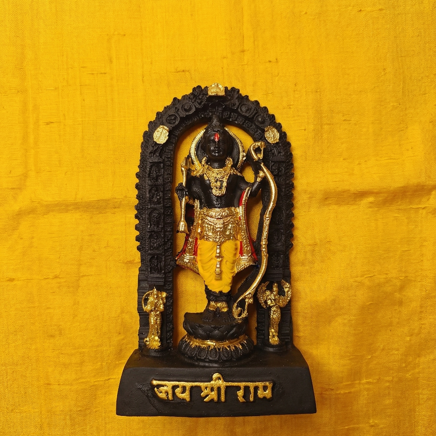 Sacred black Shri Ram Lalla murti standing inside an archway, richly adorned with golden jewelry and garments, with Jai Shri Ram inscribed on the base on yellow cloth.