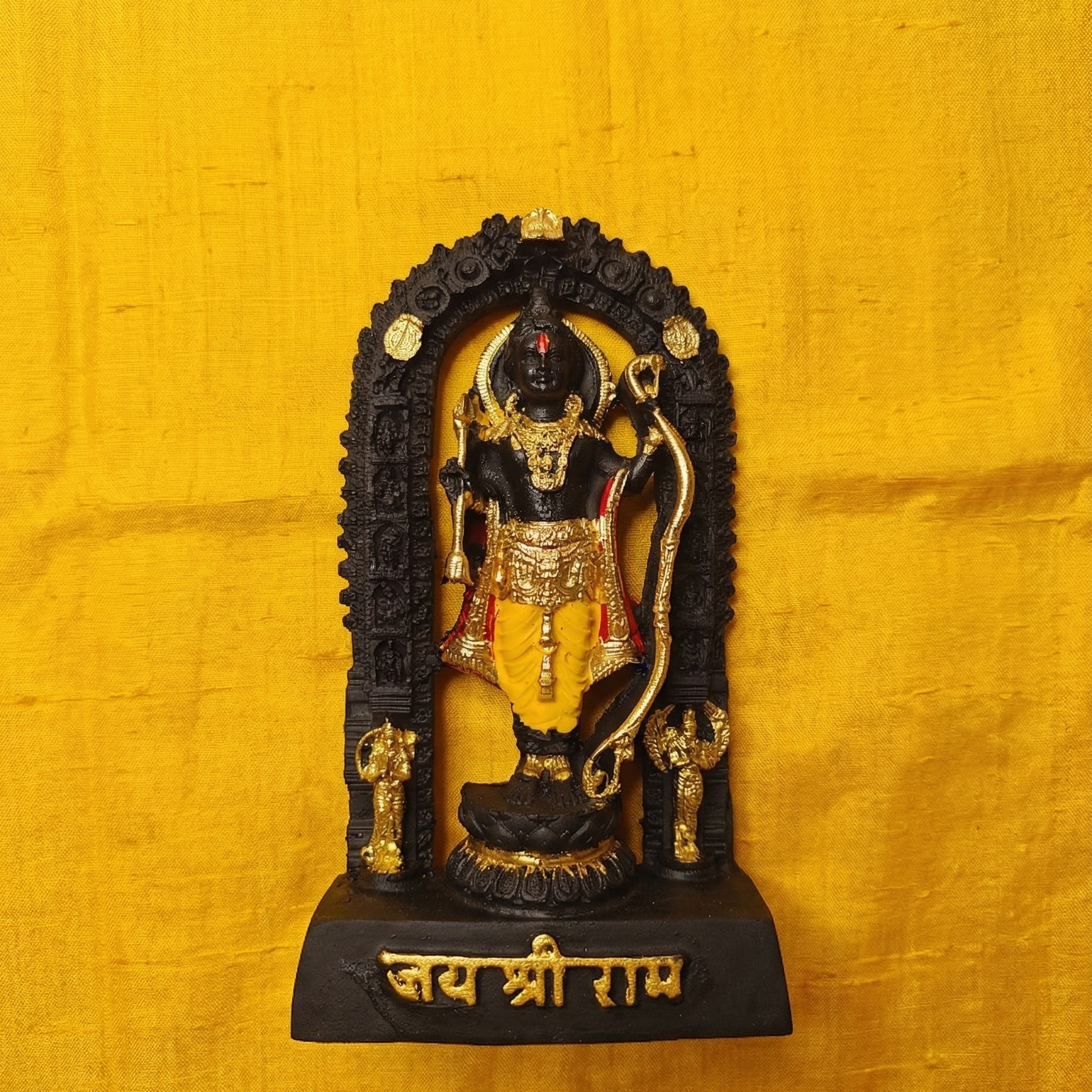 Sacred black Shri Ram Lalla murti standing inside an archway, richly adorned with golden jewelry and garments, with Jai Shri Ram inscribed on the base on yellow cloth.