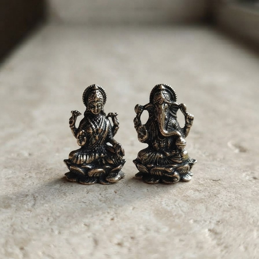 Close-up of intricately detailed miniature metal Laxmi and Ganesha idols with a dark antique finish, sitting side-by-side on a light speckled stone surface.