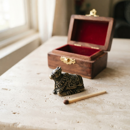 A miniature metal Nandi figurine with an antique finish sitting next to a single wooden matchstick for scale on a light stone surface, with an open wooden box in the background.
