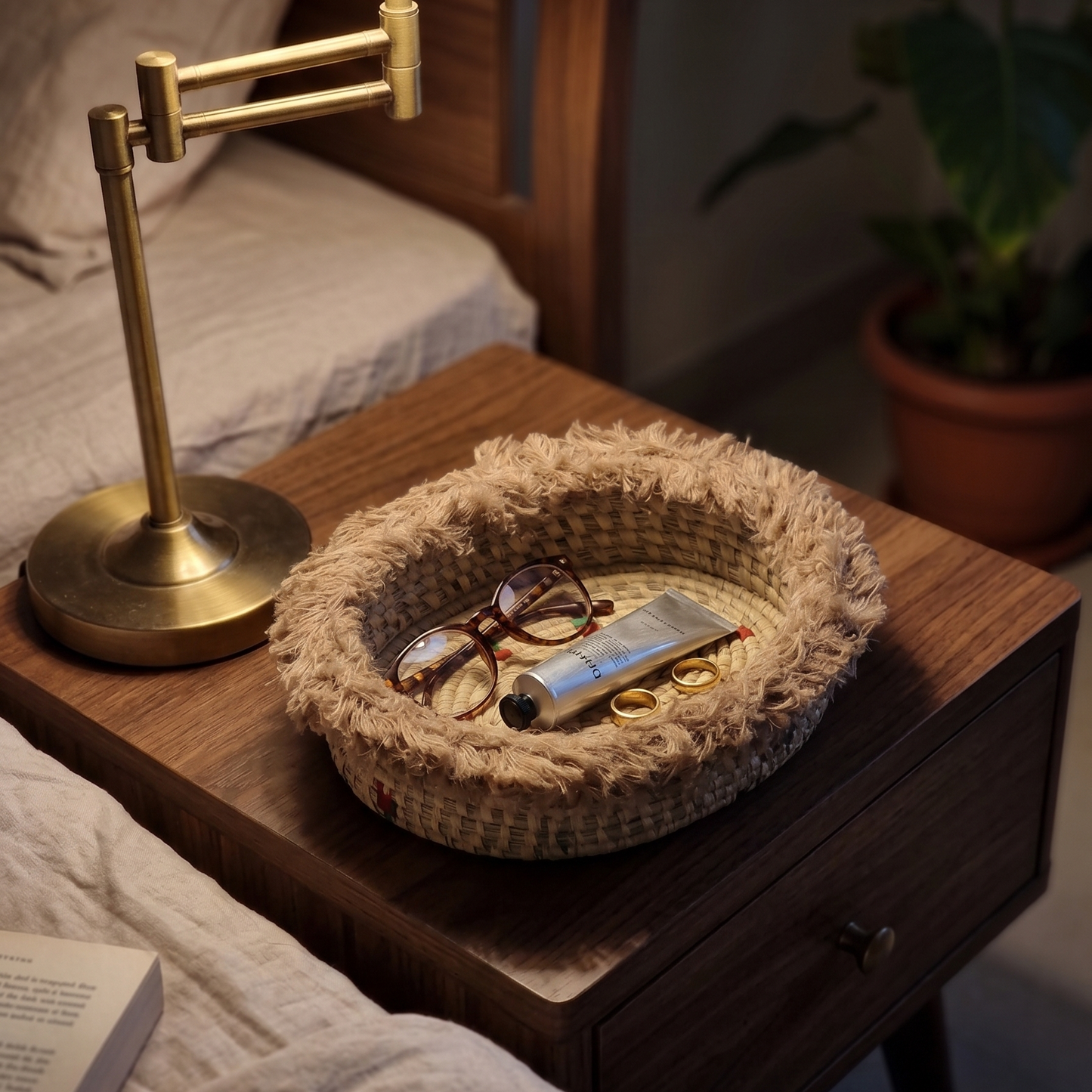 An oval, fringed Odia Heritage sabai grass basket used as a nightstand catch-all in a dimly lit, cozy bedroom. Resting on a dark walnut mid-century bedside table under a warm brass lamp, the basket holds reading glasses, a tube of hand cream, and two gold rings.