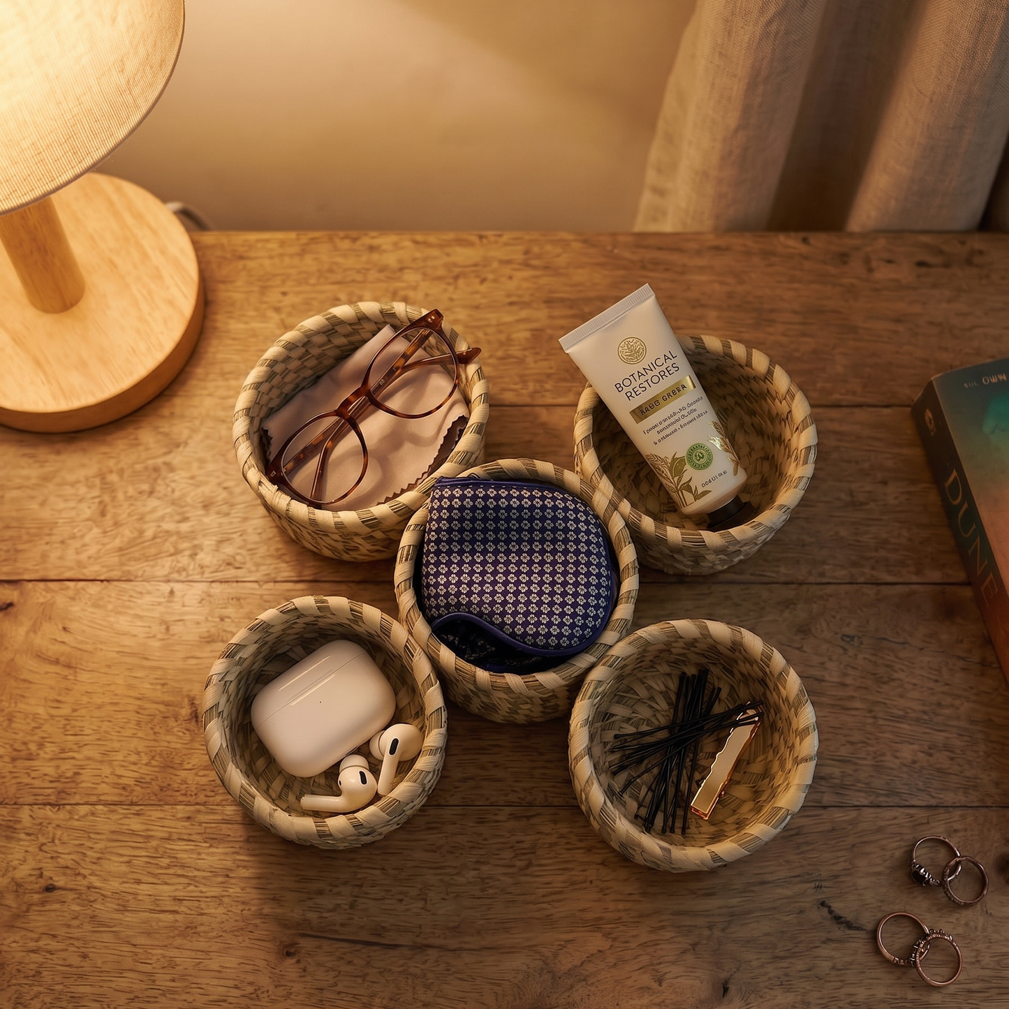 Five small, round woven baskets arranged in a cluster on a warm wooden surface under soft lighting. Each basket holds different small everyday items: one contains eyeglasses resting on a cloth, another holds a tube of botanical hand cream, a third has a white wireless earbud case with loose earbuds, the center one holds a blue patterned eye mask or pouch, and the last contains bobby pins and a hair clip.