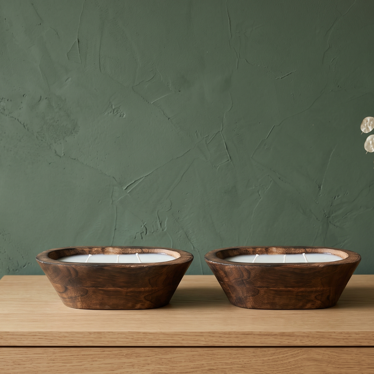 A duo set of zero-waste wooden dough bowl candles with unlit wicks, styled as organic modern decor on a light oak dresser against a textured sage green wall next to dried botanicals.
