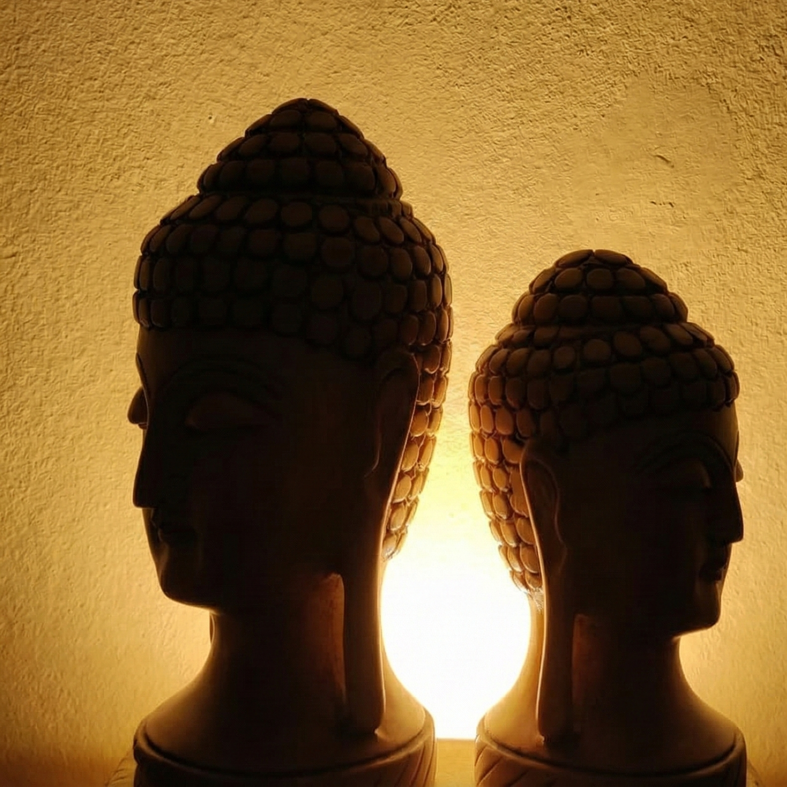 Silhouette of two wooden Buddha head statues against a warm amber light. Peaceful spiritual decor perfect for creating a calming atmosphere in bedrooms or living rooms.