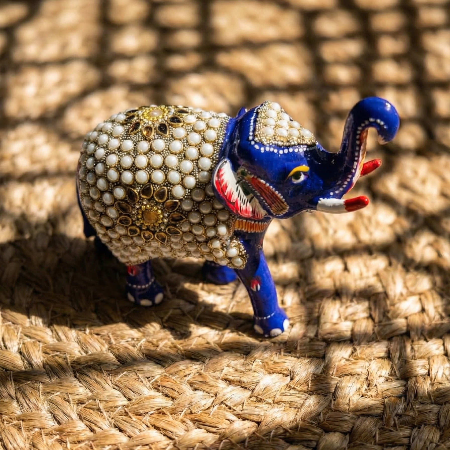 High-angle close-up of a blue Meenakari elephant statue placed on a woven mat, highlighting the intricate white pearl and kundan craftsmanship on the back.